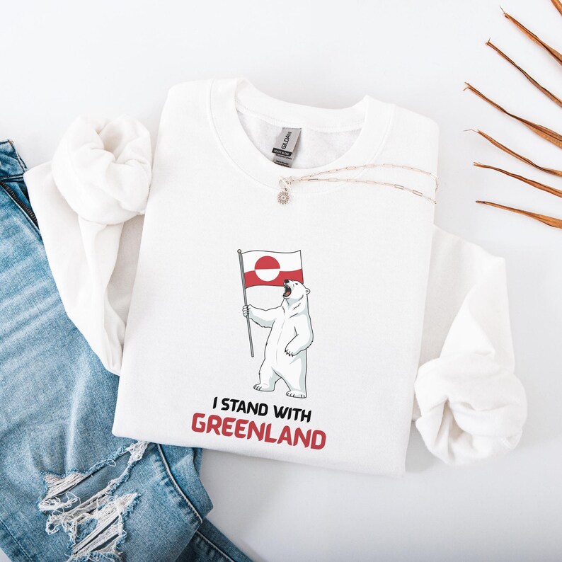 May include: White sweatshirt with a cartoon polar bear holding the Greenland flag. The text "I STAND WITH GREENLAND" is printed in red below the bear. The sweatshirt is folded on a white surface.