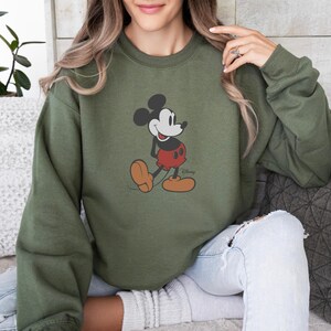 May include: An olive green sweatshirt with a Mickey Mouse graphic. The cartoon character is shown in red shorts, yellow shoes, and black ears. The Disney logo is visible on the bottom right of the graphic.