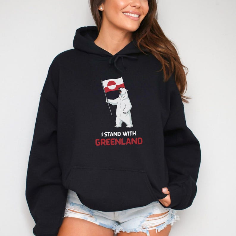 May include: Black hooded sweatshirt with a white polar bear holding a Greenland flag. The bear is in front of the text "I STAND WITH GREENLAND" in red and white. The hoodie has a drawstring hood and a front pocket.