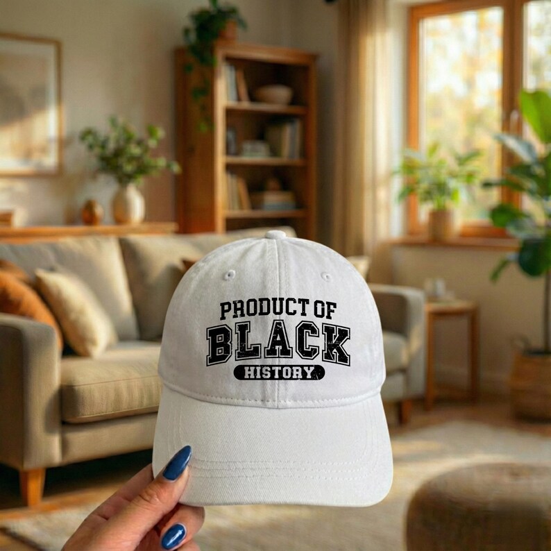 Pu&ograve; includere: Cappellino da baseball bianco con la scritta "PRODUCT OF BLACK HISTORY" in lettere nere. Il cappellino ha una visiera curva e un classico design a sei pannelli. Il cappellino &egrave; tenuto da una persona.