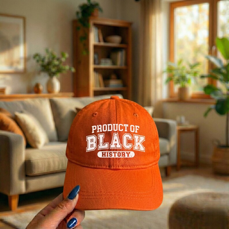 Pu&ograve; includere: Un cappellino da baseball arancione con la scritta bianca "PRODUCT OF BLACK HISTORY". Il cappellino &egrave; tenuto davanti a uno sfondo sfocato di un soggiorno con un divano e delle piante.