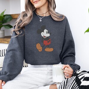 May include: Charcoal grey sweatshirt with a Mickey Mouse design. The cartoon mouse is shown in a walking pose, wearing red shorts and brown shoes. A casual, comfortable style of sweatshirt.