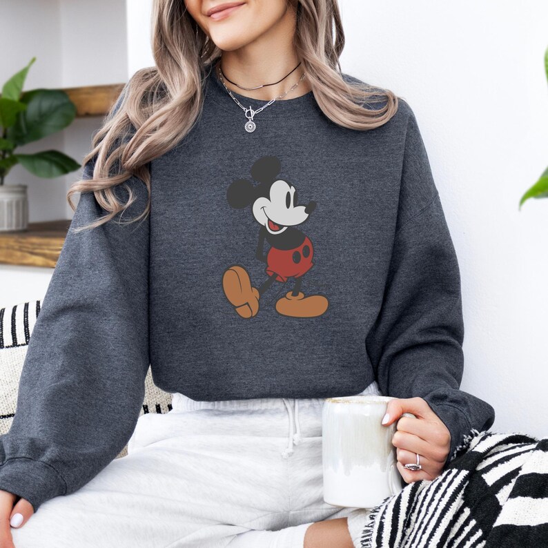 May include: Charcoal grey sweatshirt with a Mickey Mouse design. The cartoon mouse is shown in a walking pose, wearing red shorts and brown shoes. A casual, comfortable style of sweatshirt.