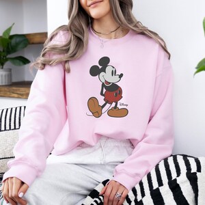 May include: A pale pink sweatshirt with a Mickey Mouse graphic. The cartoon character is shown in his classic pose, wearing red shorts and brown shoes. The Disney logo is visible. This sweatshirt is a comfortable and stylish piece of apparel.