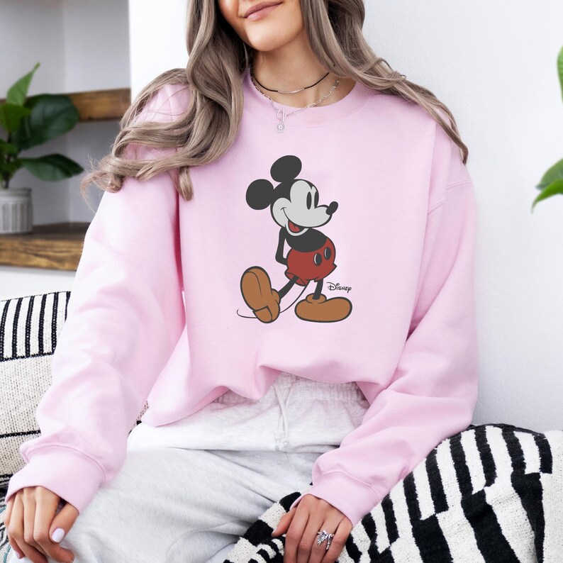 May include: A pale pink sweatshirt with a Mickey Mouse graphic. The cartoon character is shown in his classic pose, wearing red shorts and brown shoes. The Disney logo is visible. This sweatshirt is a comfortable and stylish piece of apparel.