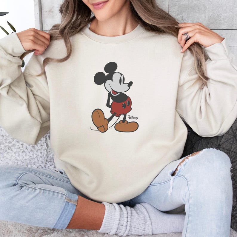 May include: A cream-coloured sweatshirt with a Mickey Mouse graphic. The cartoon character is in black, red, and brown, with the Disney logo. The long-sleeved top is a casual style.