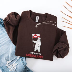 May include: A dark brown sweatshirt with a polar bear graphic holding the Greenland flag. The text "I STAND WITH GREENLAND" is printed below the bear in red and white. A silver necklace is draped across the sweatshirt.