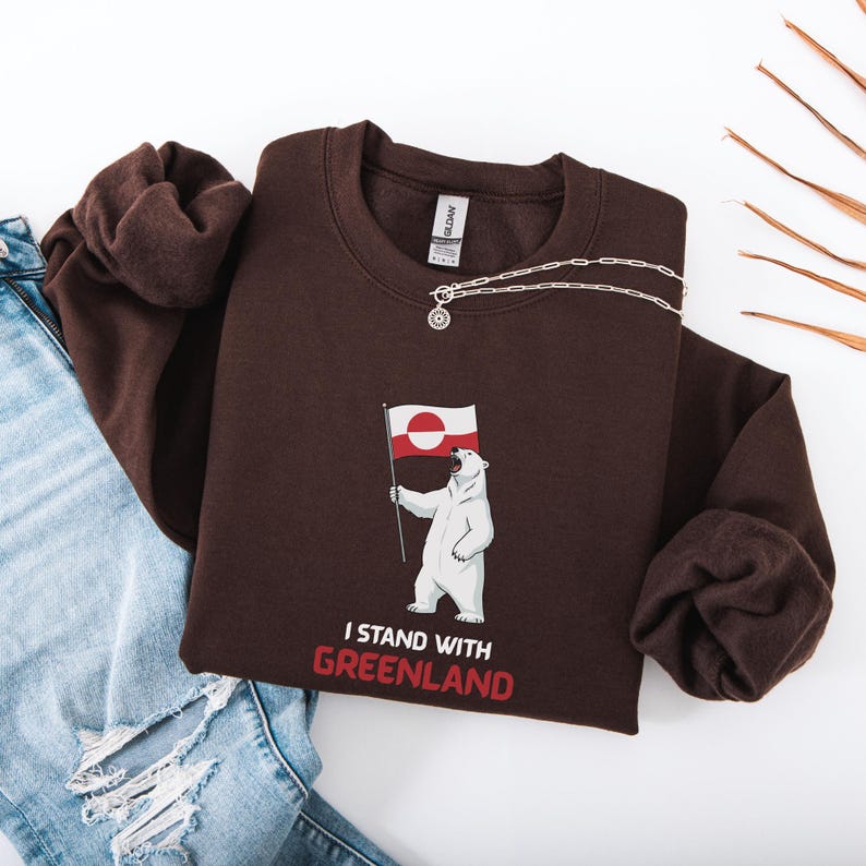 May include: A dark brown sweatshirt with a polar bear graphic holding the Greenland flag. The text "I STAND WITH GREENLAND" is printed below the bear in red and white. A silver necklace is draped across the sweatshirt.