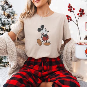 May include: A beige t-shirt with a Mickey Mouse graphic in black, red, and brown. The person is wearing red and black plaid pyjama bottoms and a cream cardigan. A Christmas tree and a mug with a Santa Claus design are in the background.