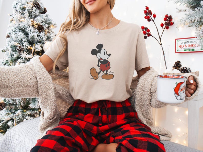 May include: A beige t-shirt with a Mickey Mouse graphic in black, red, and brown. The person is wearing red and black plaid pyjama bottoms and a cream cardigan. A Christmas tree and a mug with a Santa Claus design are in the background.