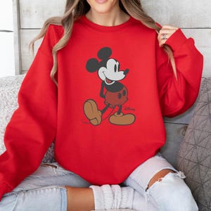May include: A red sweatshirt with a Mickey Mouse graphic. The cartoon character is shown in his classic pose, with black ears, a white face, red shorts, and brown shoes. The Disney logo is visible at the bottom right.