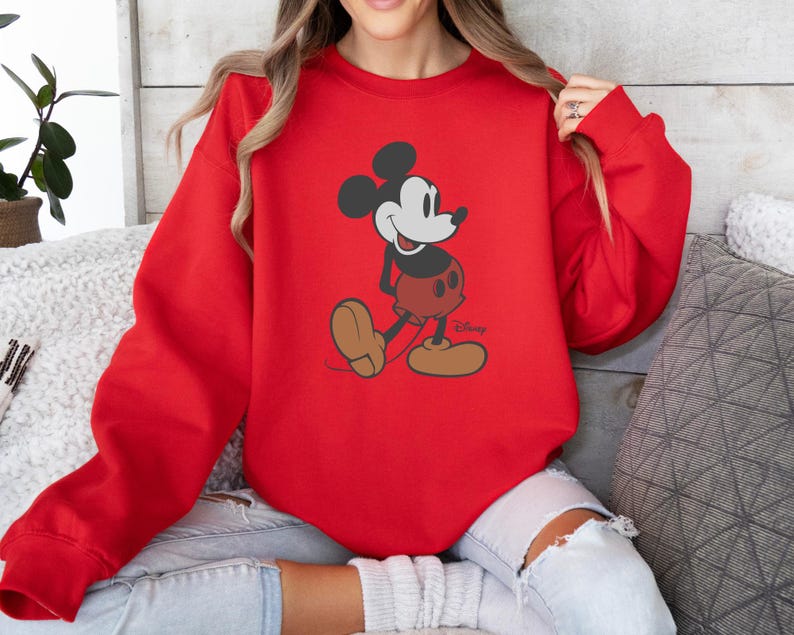 May include: A red sweatshirt with a Mickey Mouse graphic. The cartoon character is shown in his classic pose, with black ears, a white face, red shorts, and brown shoes. The Disney logo is visible at the bottom right.