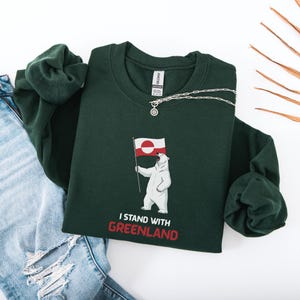 May include: A dark green sweatshirt with a polar bear graphic holding a Greenland flag. The text "I STAND WITH GREENLAND" is printed in red. A silver necklace is draped across the sweatshirt. The sweatshirt is folded.