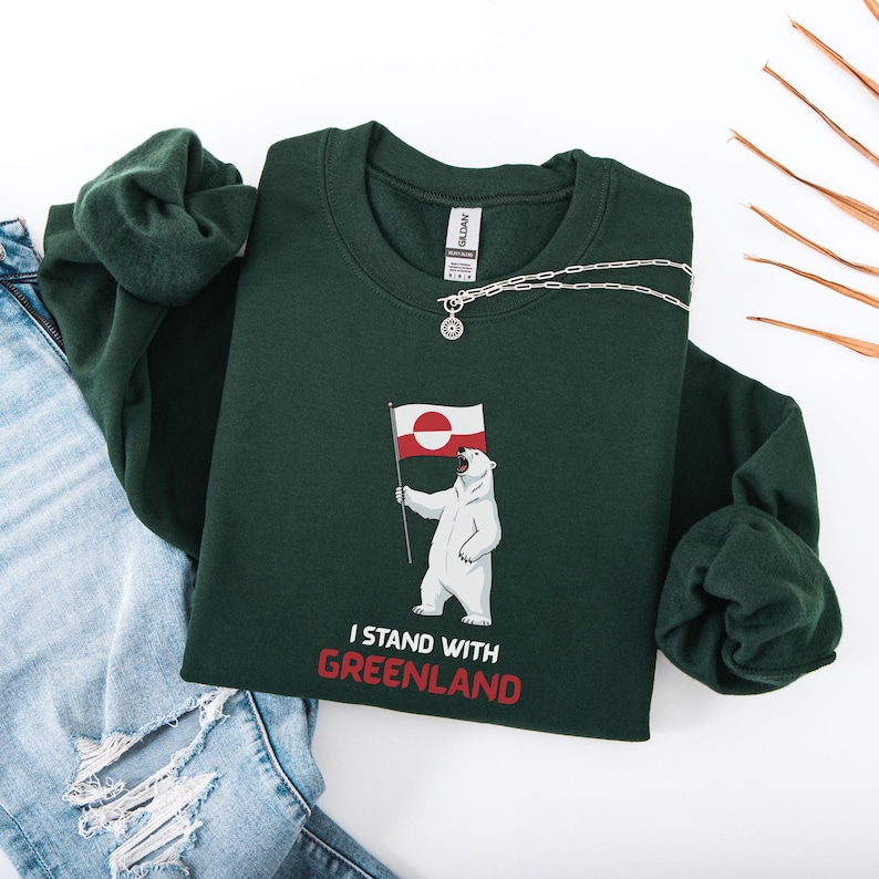 May include: A dark green sweatshirt with a polar bear graphic holding a Greenland flag. The text "I STAND WITH GREENLAND" is printed in red. A silver necklace is draped across the sweatshirt. The sweatshirt is folded.