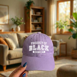 Pu&ograve; includere: Un cappellino da baseball lavanda con la scritta "PRODUCT OF BLACK HISTORY" in lettere bianche. Il cappellino ha una visiera curva e un classico design a sei pannelli. Lo sfondo &egrave; un soggiorno sfocato.