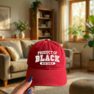 Pu&ograve; includere: Cappellino da baseball rosso con la scritta bianca "PRODUCT OF BLACK HISTORY". Il cappellino ha una visiera curva ed &egrave; realizzato in un tessuto morbido. Lo sfondo mostra un soggiorno con un divano e mobili in legno.