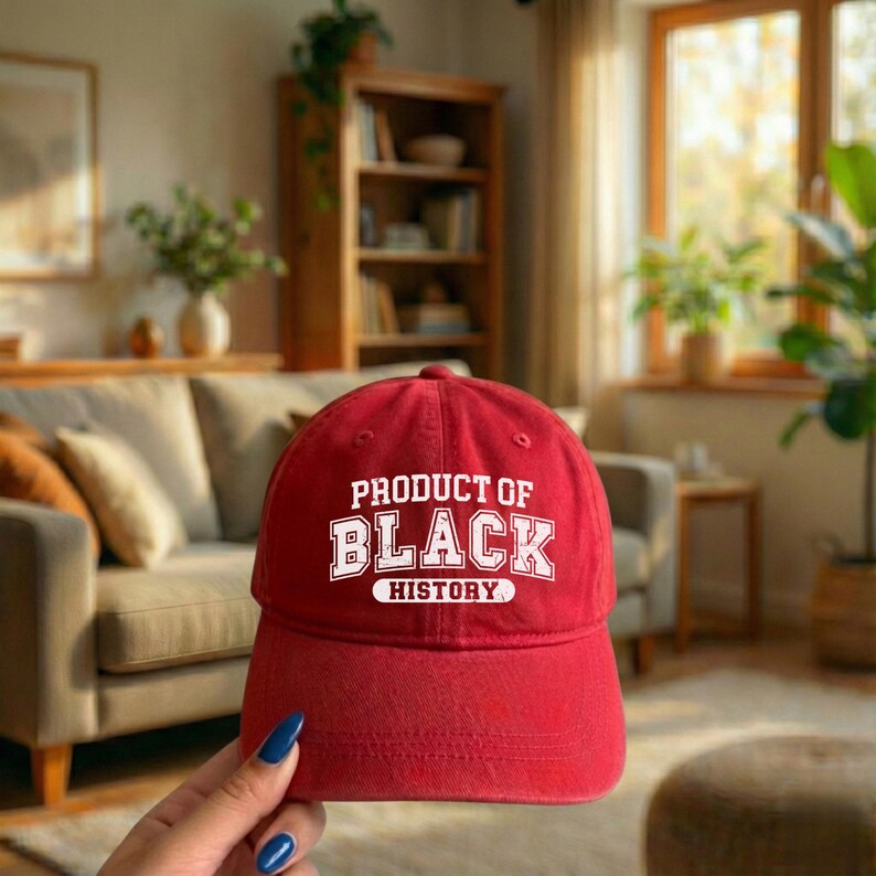 Pu&ograve; includere: Cappellino da baseball rosso con la scritta bianca "PRODUCT OF BLACK HISTORY". Il cappellino ha una visiera curva ed &egrave; realizzato in un tessuto morbido. Lo sfondo mostra un soggiorno con un divano e mobili in legno.