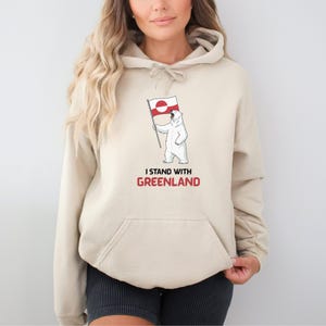 May include: Beige hoodie featuring a polar bear holding a Greenland flag, with the text "I STAND WITH GREENLAND" printed below. The hoodie has a drawstring hood and a front pocket. The design is in red and white.