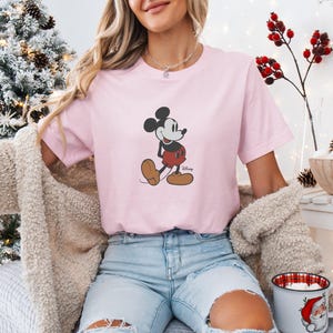 May include: A pale pink t-shirt with a Mickey Mouse design. The cartoon character is shown in his classic pose, wearing red shorts and brown shoes. The shirt is part of a Disney collection.