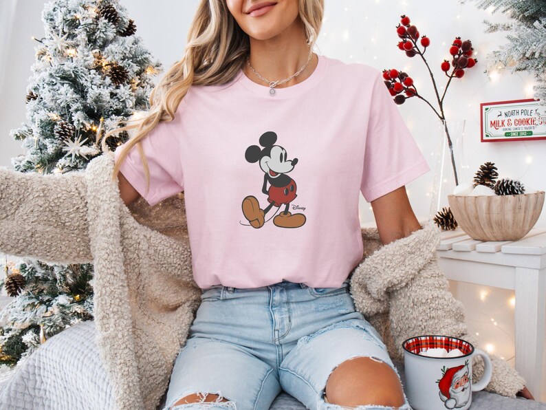 May include: A pale pink t-shirt with a Mickey Mouse design. The cartoon character is shown in his classic pose, wearing red shorts and brown shoes. The shirt is part of a Disney collection.
