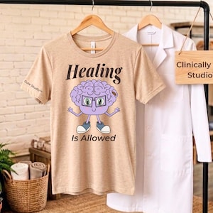 May include: Beige t-shirt with the words "Healing Is Allowed" and a cartoon brain character wearing glasses. The shirt is hanging on a wooden hanger. A white lab coat hangs next to it. A sign reads "Clinically Cute Studio™".