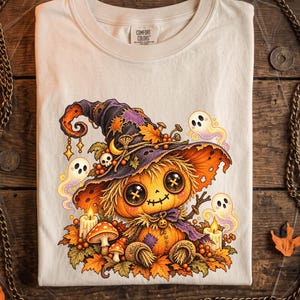 May include: An ivory t-shirt featuring a graphic print of a pumpkin character with button eyes wearing a large witch hat. The design includes small ghosts, lit candles, mushrooms, and autumn leaves. The shirt has a crew neckline and a label.