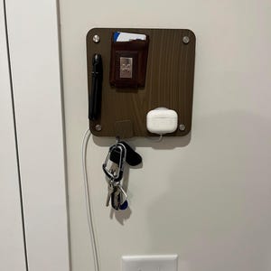 Könnte beinhalten: Ein brauner Wandorganizer aus Holz mit einer Brieftasche, Schlüsseln, einer Taschenlampe und kabellosen Ohrhörern. Der Organizer ist an einer weißen Wand montiert, darunter ein Lichtschalter.