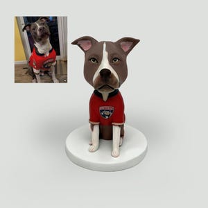 Può includere: Una statuetta bobblehead personalizzata di un cane pitbull marrone e bianco che indossa una maglia rossa con un logo a forma di pantera sul petto. Il cane ha orecchie dritte e una striscia bianca sul muso. La statuetta è su una base circolare.