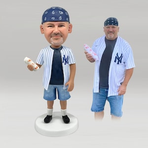 May include: A custom bobblehead figurine depicting a person wearing a dark blue bandana, a white pinstriped baseball jersey with a navy blue NY logo, a dark t-shirt, blue denim shorts, white socks, and black shoes, holding a white wrapped food item.