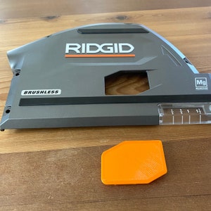 May include: A gray Ridgid power tool with a black and orange label that says "Ridgid" and "Brushless". The tool has a clear ruler on the side. A small orange plastic piece is in the foreground.