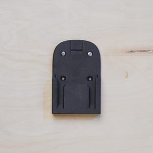 Op de afbeelding: Zwarte plastic wandbevestiging met twee schroefgaten bovenaan en twee onderaan. De bevestiging heeft een rechthoekige opening in het midden.