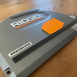 May include: A gray power tool with the word "RIDGID" in white lettering and the word "BRUSHLESS" in black lettering. The tool has an orange hexagonal button.