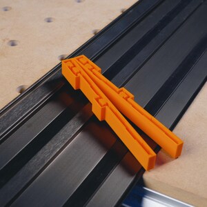 May include: Two orange plastic T-track fence clamps for woodworking. The clamps are placed on a black T-track.