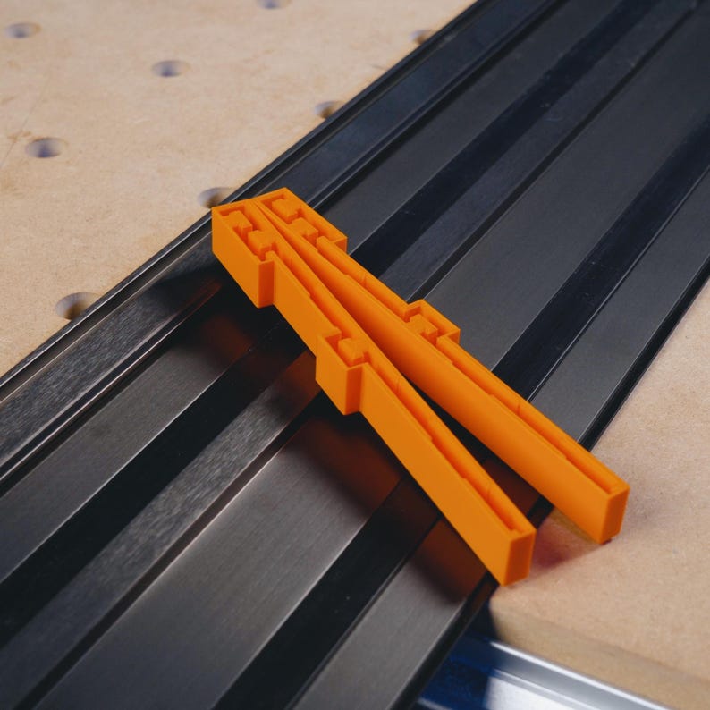 May include: Two orange plastic T-track fence clamps for woodworking. The clamps are placed on a black T-track.