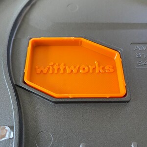 May include: Close-up of an orange plastic label with the word "Wittworks" embossed on it. The label is attached to a gray surface.