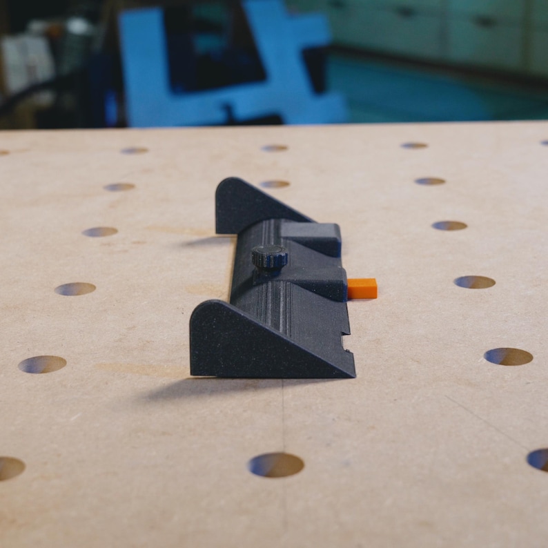 May include: A black plastic woodworking tool with a knob and an orange tab. The tool is on a light brown surface with holes.