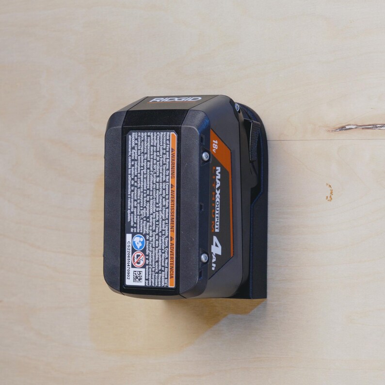 Op de afbeelding: Een zwarte en oranje 18v 4Ah MAX Lithium batterij voor een Ridgid powertool. De batterij is gemonteerd op een zwarte wandbevestiging.