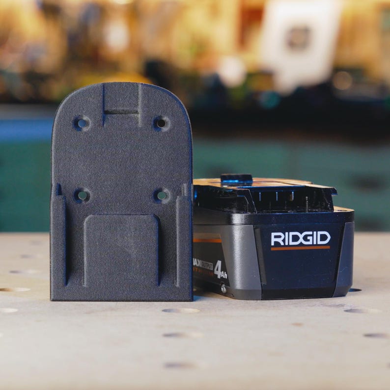 Op de afbeelding: Een zwarte plastic wandbevestiging voor een Ridgid 4Ah batterij. De bevestiging heeft vier schroefgaten en een rechthoekige opening voor de batterij.