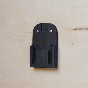Op de afbeelding: Zwarte plastic wandbevestiging voor een batterijlader. De beugel heeft twee schroefgaten bovenaan en twee onderaan.