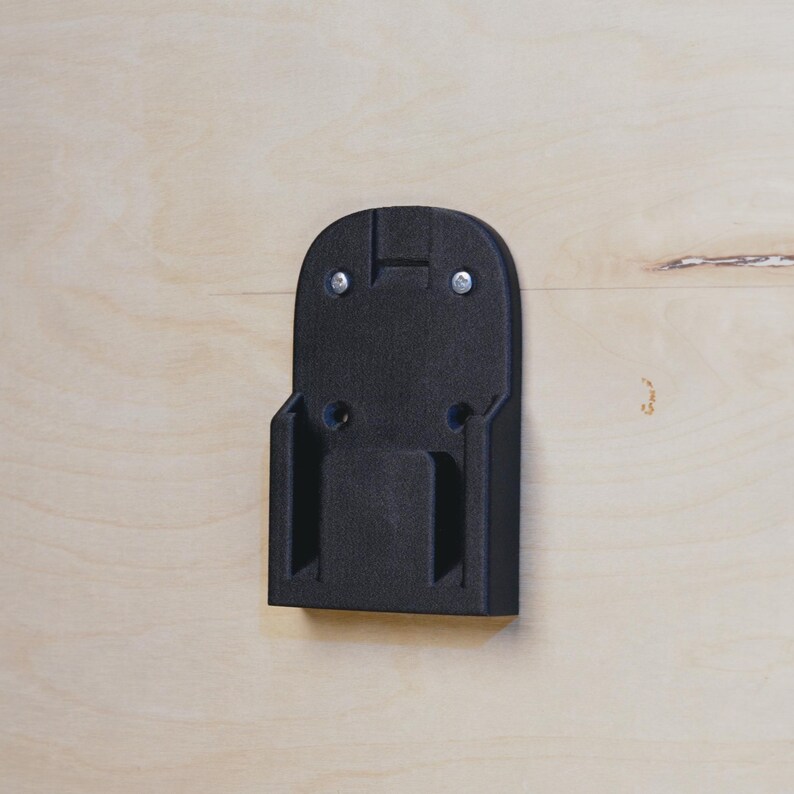 Op de afbeelding: Zwarte plastic wandbevestiging voor een batterijlader. De beugel heeft twee schroefgaten bovenaan en twee onderaan.