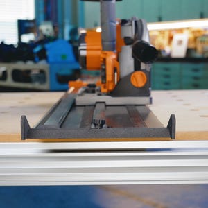 May include: A close-up of a black and orange power tool with a black and silver track system. The tool is mounted on a wooden surface.