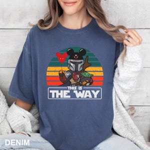 May include: Denim blue t-shirt featuring a graphic of a Mandalorian character with a Mickey Mouse head, holding a red heart balloon, and Baby Yoda. The text "THIS IS THE WAY" is displayed in a white rectangle against a sunset background.