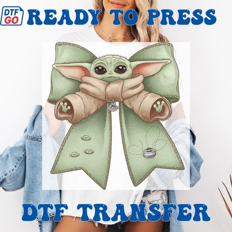 Puede incluir: Una transferencia DTF con un lazo verde y un personaje de dibujos animados en el centro. El personaje tiene ojos grandes y est&aacute; envuelto en una manta marr&oacute;n. El lazo tiene tres peque&ntilde;os dise&ntilde;os de nave espacial y el texto "READY TO PRESS" y "DTF TRANSFER" en azul.