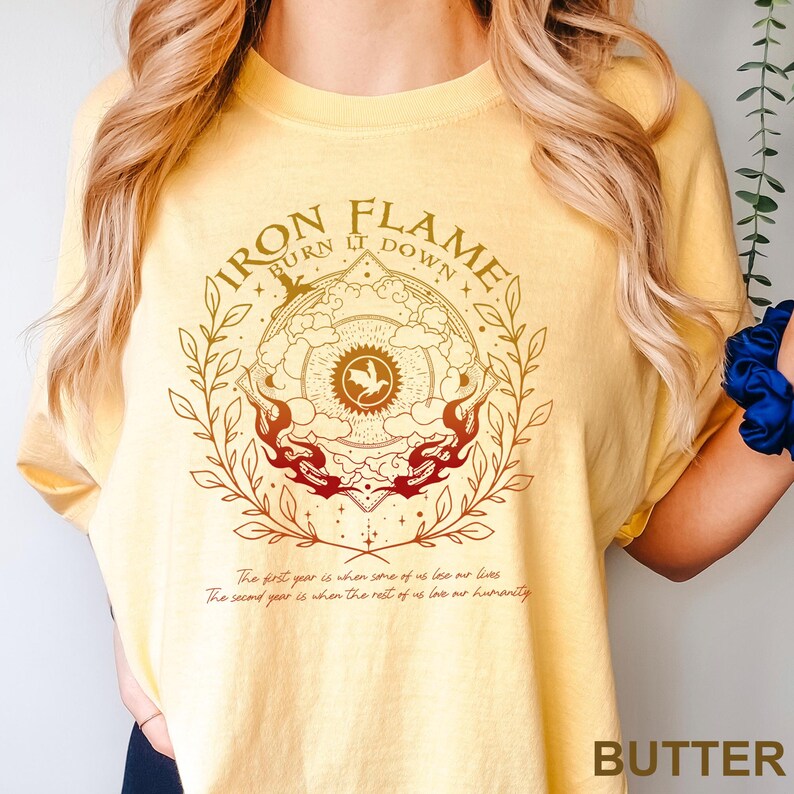 May include: Butter-colored t-shirt with a circular graphic featuring the text "IRON FLAME BURN IT DOWN." The design includes a central emblem with a bird, surrounded by flames and botanical elements. Below the graphic are two lines of text.
