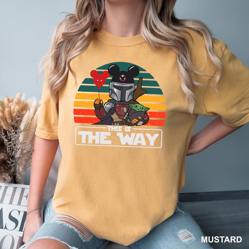May include: Mustard-colored t-shirt featuring a graphic of The Mandalorian with Mickey Mouse ears holding a heart-shaped balloon, Grogu, and the text "THIS IS THE WAY" against a sunset backdrop.