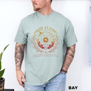 May include: A light green t-shirt with a circular graphic that reads "IRON FLAME BURN IT DOWN". The graphic features a central design with flames and a floral wreath. The text "BAY" is at the bottom right.