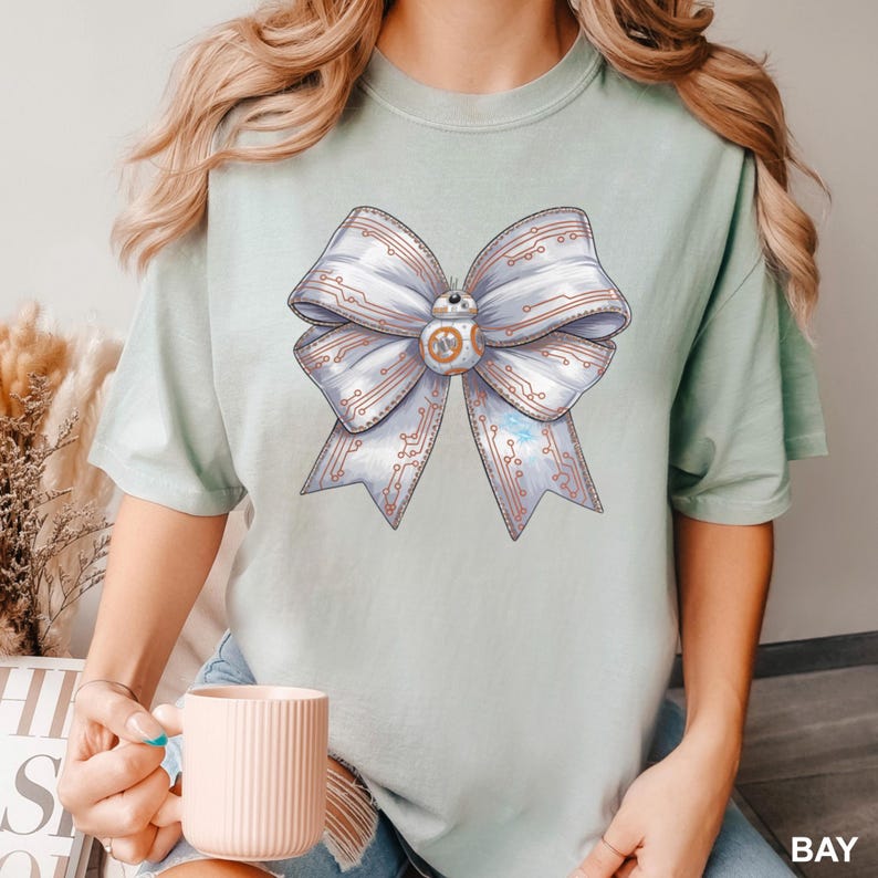 Puede incluir: Camiseta verde claro con un gran dise&ntilde;o de lazo con patrones de placa de circuito y un droide BB-8 en el centro. El lazo es blanco con detalles en rojo y azul. La camiseta es de color s&oacute;lido.
