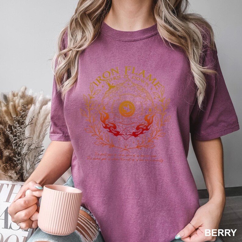 May include: Berry-colored t-shirt with a circular graphic design in shades of orange and yellow, featuring the text "IRON FLAME". The design includes floral and flame elements. The shirt is a comfortable, casual style.