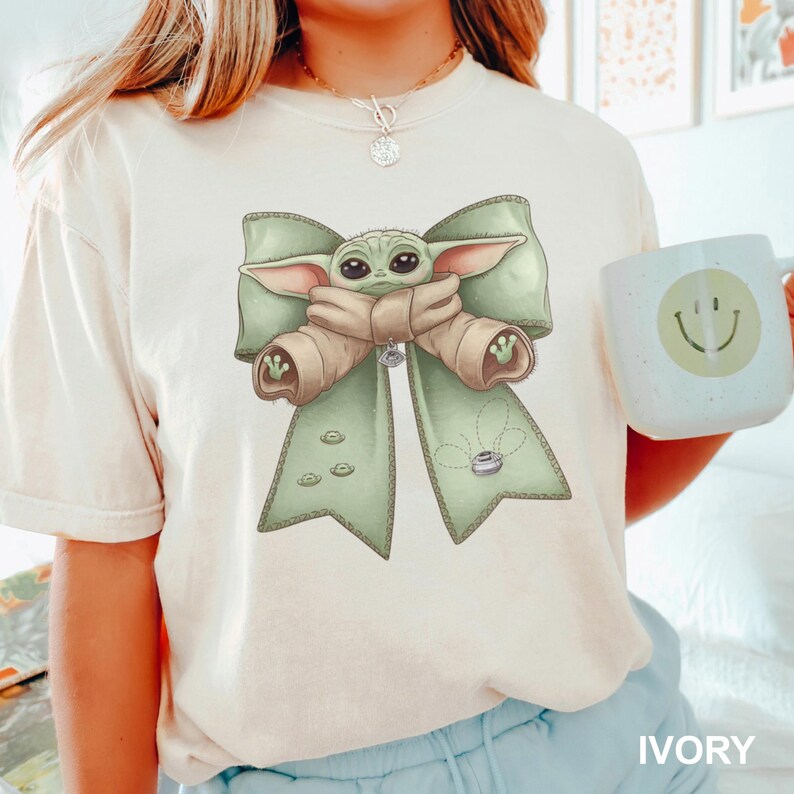 Puede incluir: Camiseta color marfil con un gr&aacute;fico de un personaje alien&iacute;gena verde con un lazo verde claro y una cinta beige. Una taza blanca con una carita sonriente se sostiene en la mano. La palabra "IVORY" est&aacute; impresa en la parte inferior.