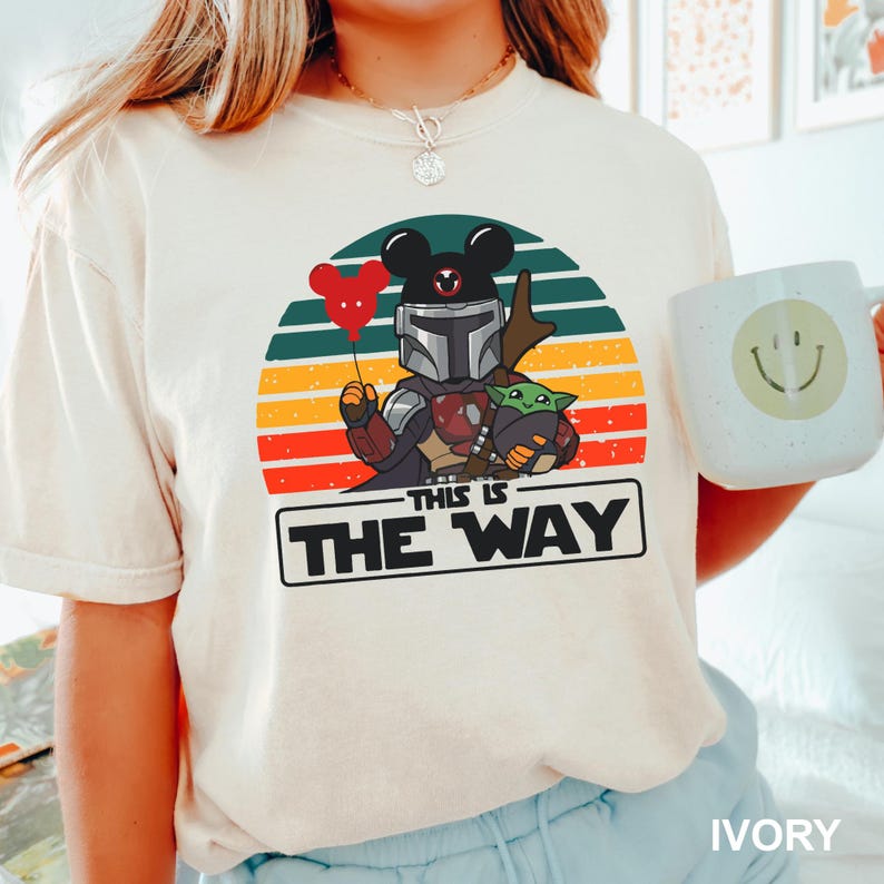 May include: Ivory-colored t-shirt featuring a graphic of a Mandalorian character with Mickey Mouse ears, holding a red heart balloon and Grogu. The design is set against a sunset backdrop with the text "THIS IS THE WAY". A white mug with a smiley face is also visible.